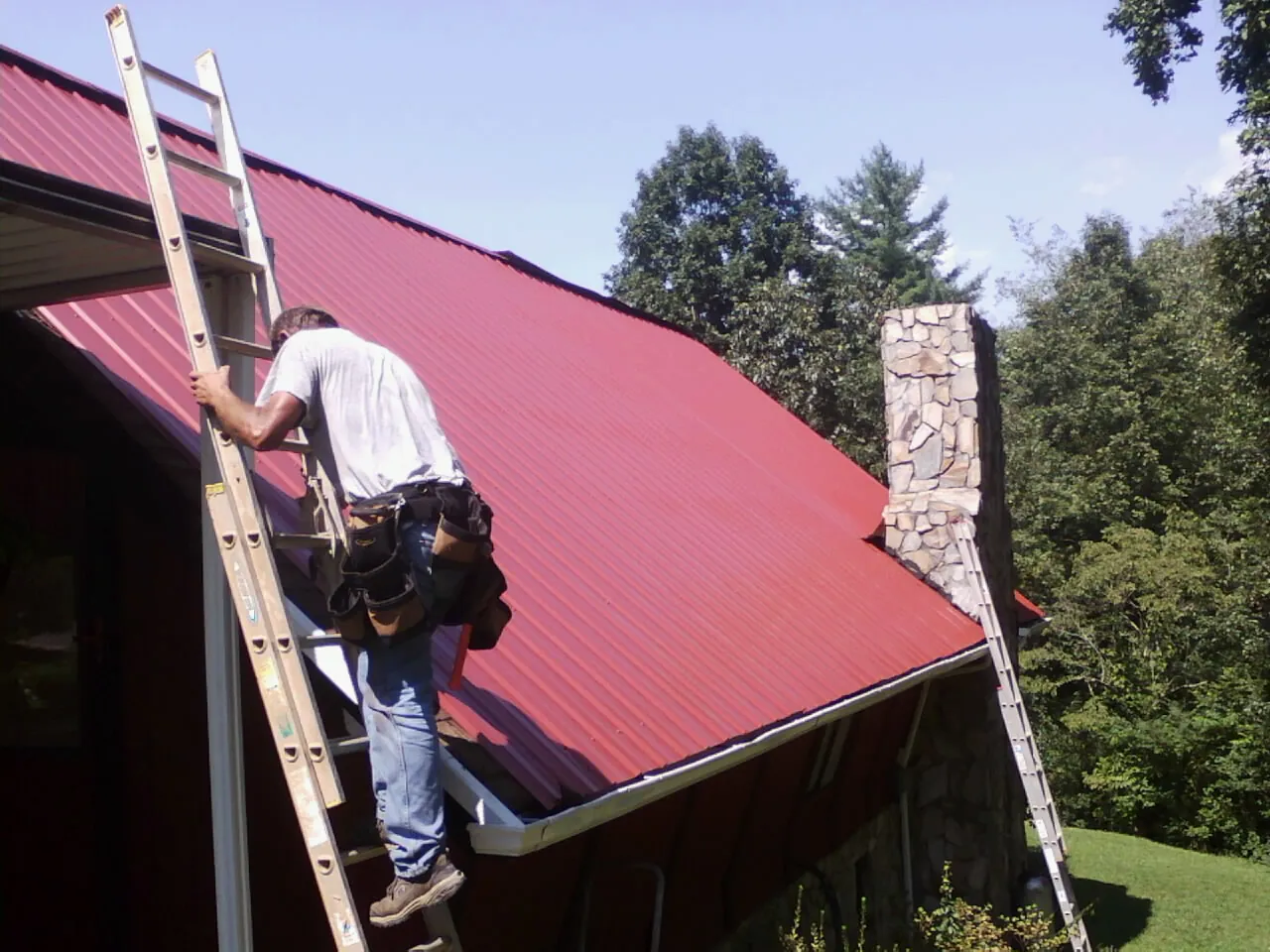 Roofer on a ladder beside a red metal roof and stone chimney — Soffit & Fascia Repair by Roofing Brothers in Greensboro